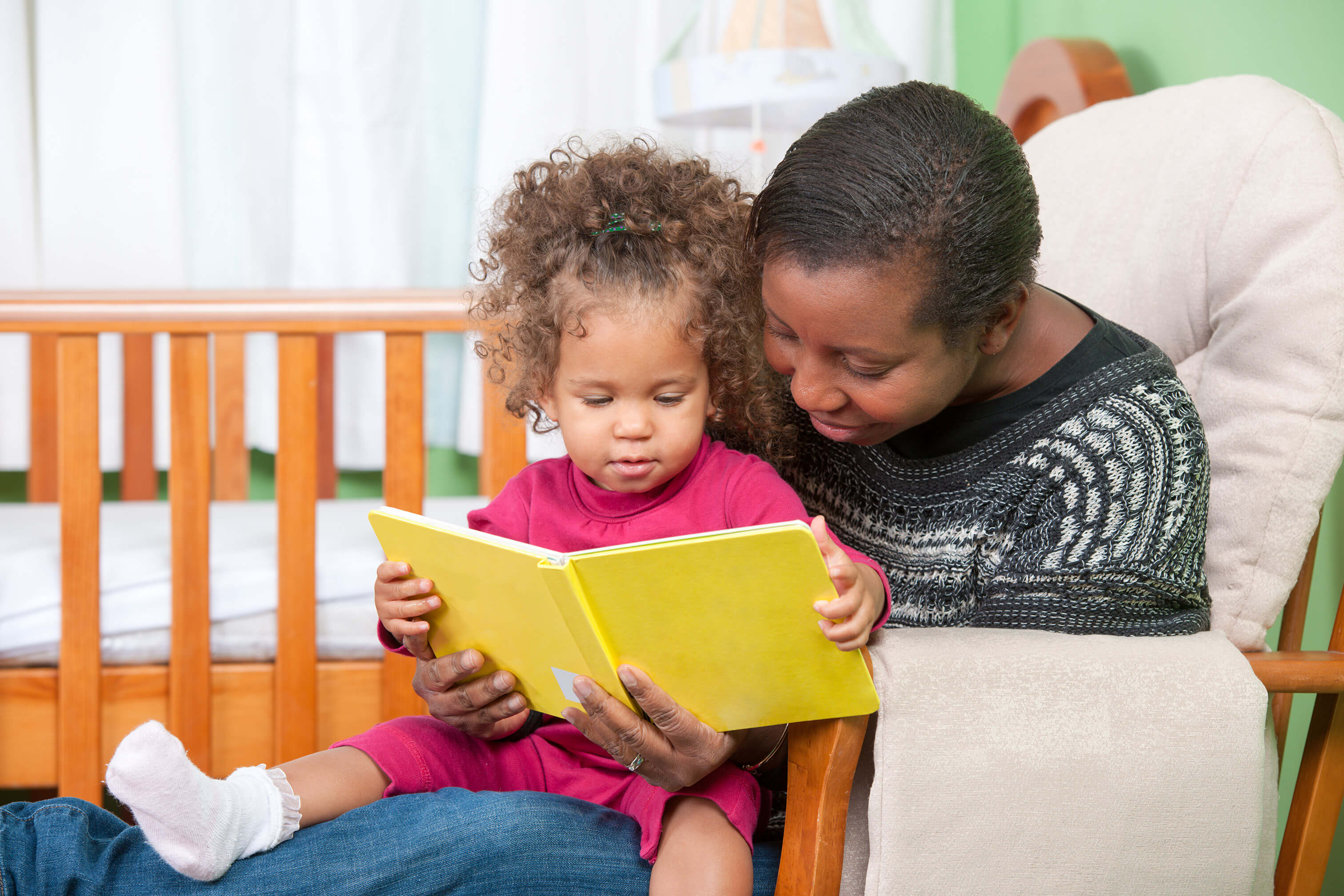 Child care provider sitting in chair reading with toddler
