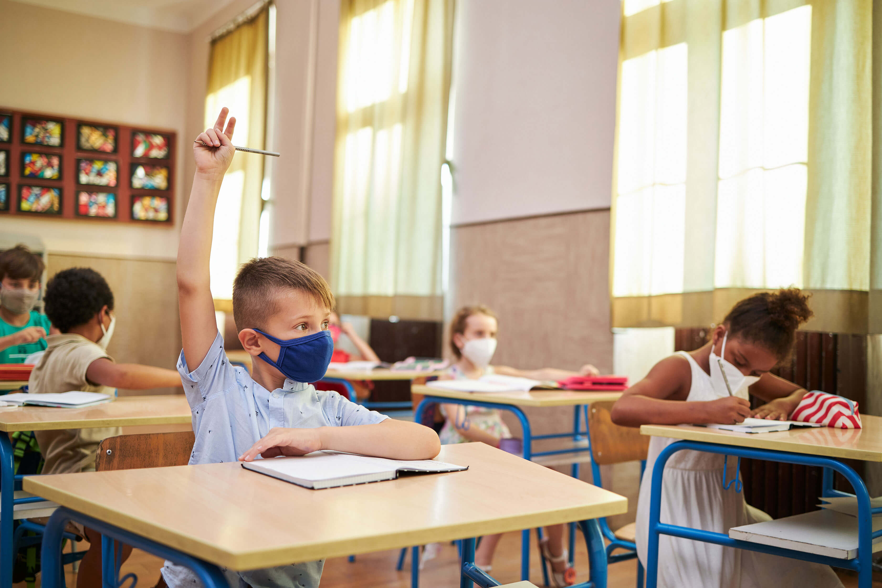 A boy raising his hand, while wearing a mask, in a classroom