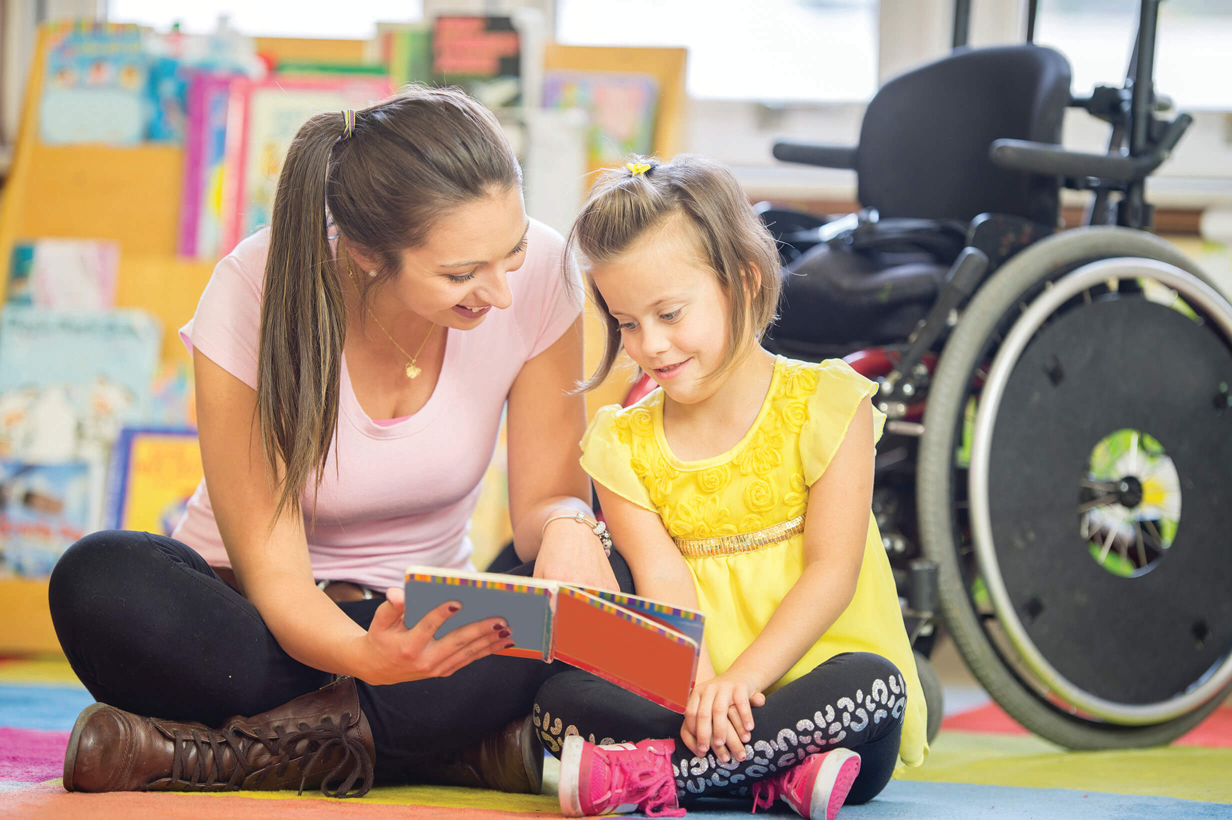 Una proveedora de cuidado infantil sentada en el suelo con un estudiante en silla de ruedas